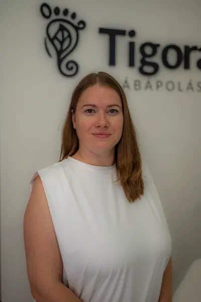 Judit, a Tigora Lábápolás gyógypedikűröse a rendelőben, a Tigora logó előtt